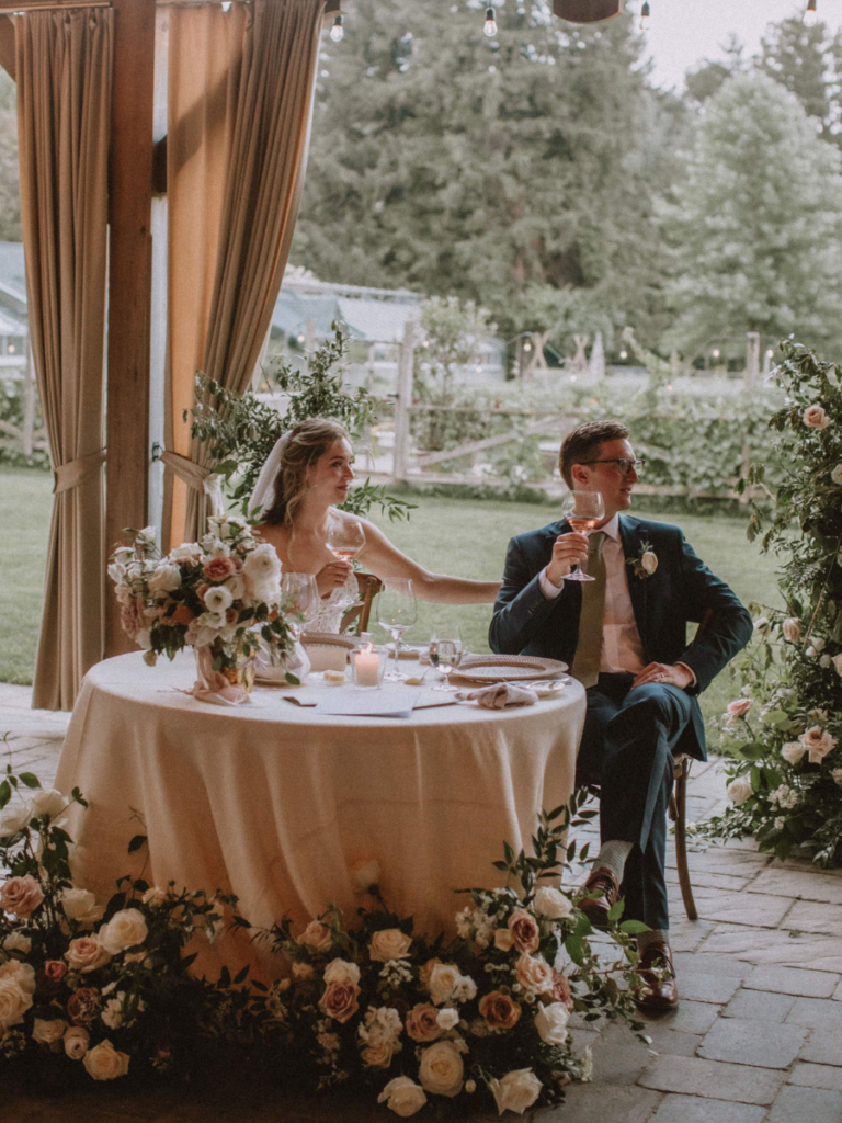 Bride & Groom sitting at their sweetheart table surrounded by florals and enjoying listening to speeches