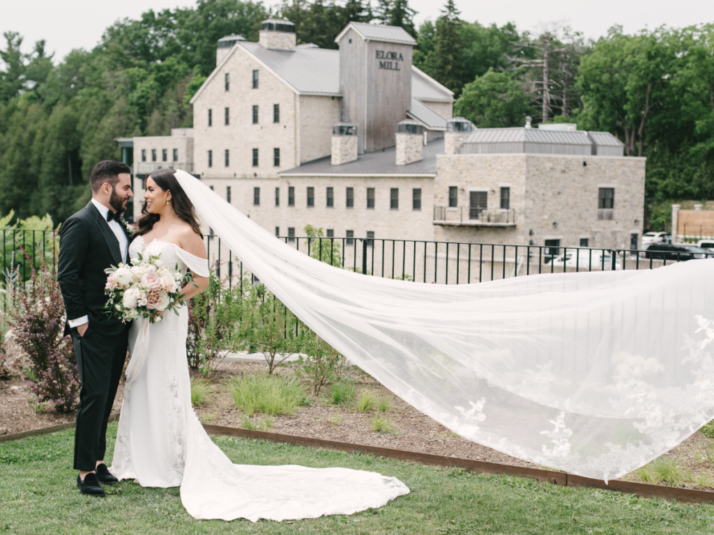 Bride & Groom in front of the Elora Mill with brides veil flowing in the wind